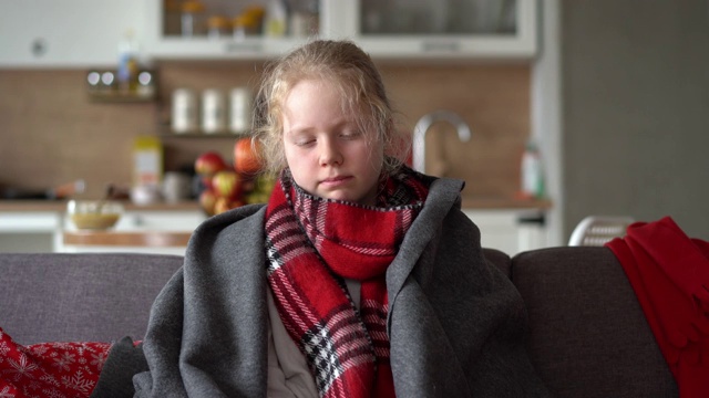 家里有个冷白人女孩。一个戴着围巾和格子图案的生病的孩子躺在公寓的沙发上，女学生用餐巾擤鼻涕视频素材