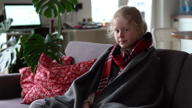 家里有个冷白人女孩。一个戴着围巾和格子图案的生病的孩子躺在公寓的沙发上，女学生用餐巾擤鼻涕视频素材