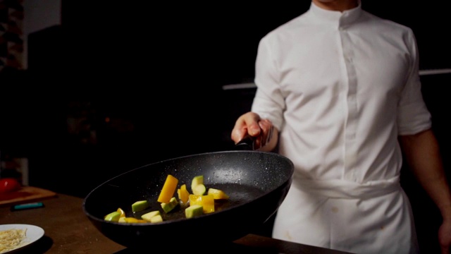 在餐厅厨房用平底锅煎炸蔬菜时，用慢镜头拍下一位面熟的年轻厨师翻炒切好的蔬菜(黄椒和西葫芦)视频素材