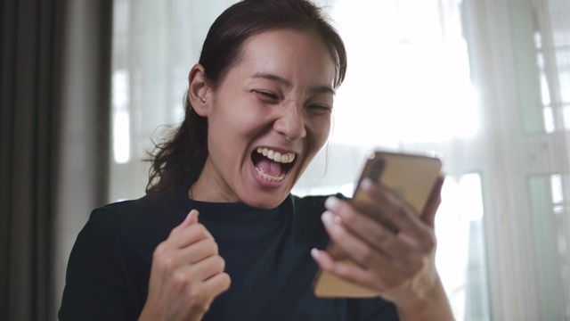 年轻的亚洲女性拿着智能手机，庆祝获奖或欢呼手机奖视频素材