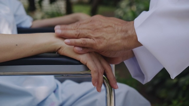 关怀的手，亚洲医生在轮椅与花园背景妇女的手。手的闭合。视频素材