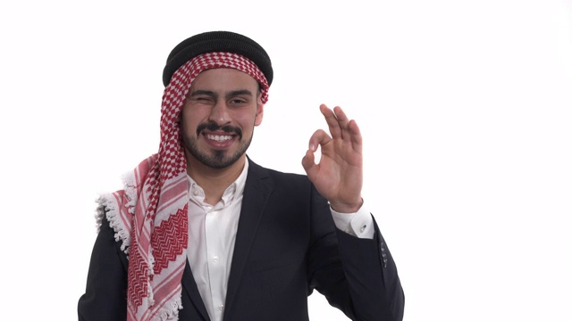 微笑的阿拉伯人在kufiya民族显示ok标志和看着相机在白色的背景视频素材