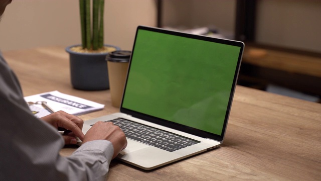 一个男人的手在笔记本电脑的绿色屏幕上工作的特写。在背景模糊和明亮的办公室里，一个人靠近另一个人，他们在讨论。视频素材