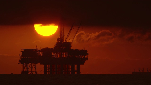 太阳从风暴云下与一个剪影的近海石油钻井平台在前景和石油(石油)油轮在背景在日落下面戏剧性的，暴风雨的天空视频下载