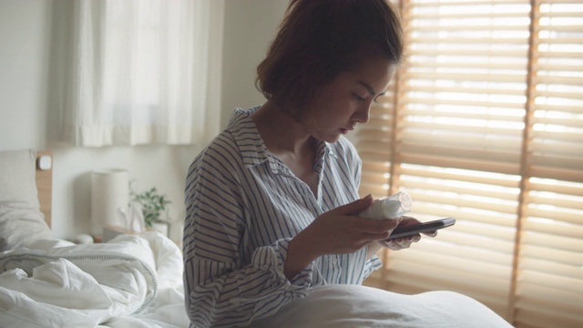 年轻的亚洲女人拿着药瓶，在卧室里讲手机视频下载