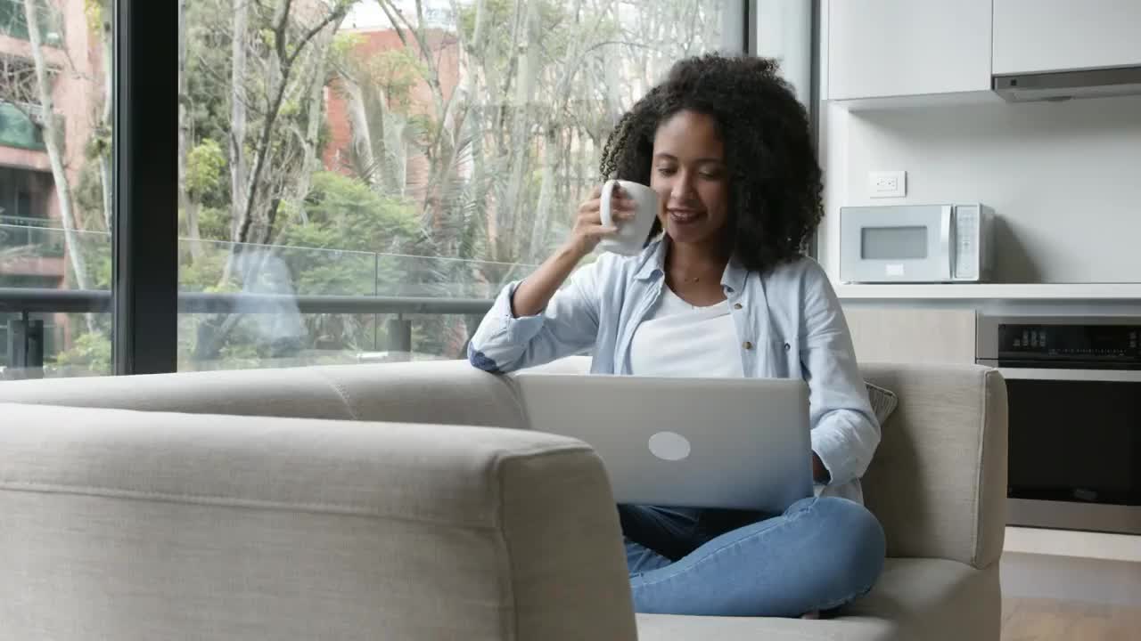 美丽的黑人女子坐在家里的沙发上，一边享受着热巧克力，一边浏览社交媒体视频素材