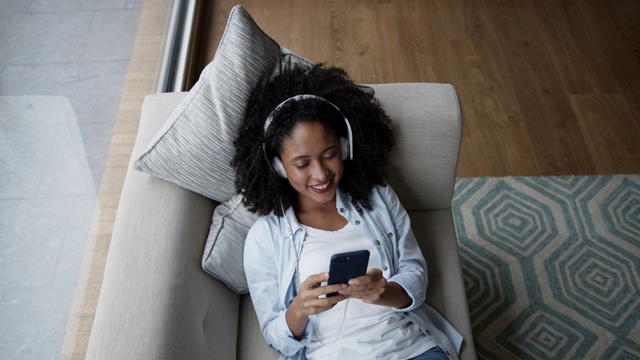 高角度的黑人妇女放松在沙发上听音乐耳机和使用智能手机看起来非常高兴视频素材