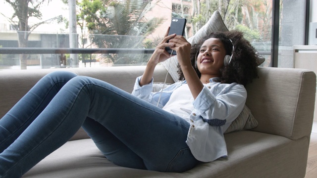 美丽的女人放松在家里的沙发上听音乐耳机，看着智能手机屏幕微笑视频素材