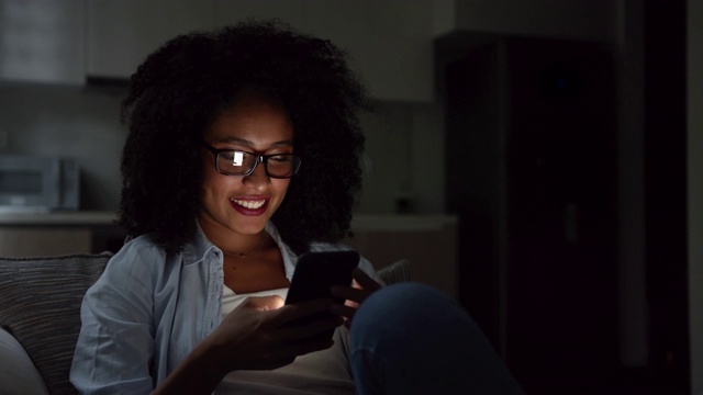 一名非裔美国女性，晚上躺在沙发上，戴着眼镜用智能手机看社交媒体视频素材