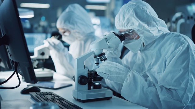 研究工厂洁净室:工程师/科学家穿着工作服和手套使用显微镜检查主板微处理器组件，开发高科技现代电子视频素材