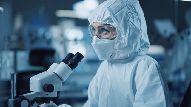 研究工厂洁净室:工程师/科学家穿着工作服和使用显微镜检查样品，为医疗和高精密电子工业开发高科技现代电子视频素材