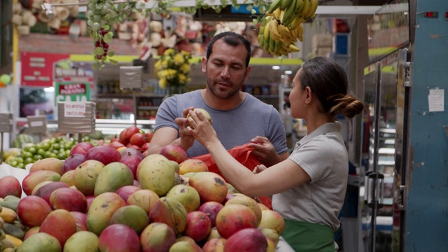 一位拉丁美洲的女售货员在农贸市场帮助顾客选择芒果，同时他把芒果放入可重复使用的袋子里视频素材
