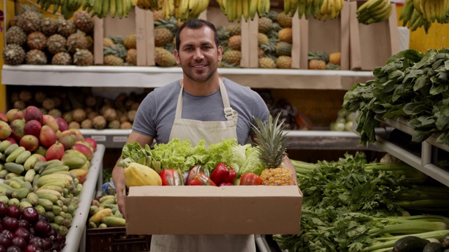 一名友善的男子正准备在一个装满新鲜水果和蔬菜的纸箱里为顾客送货，微笑着看着相机视频素材