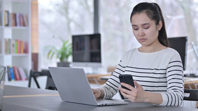 年轻的亚洲女性使用笔记本电脑在办公室使用智能手机视频素材