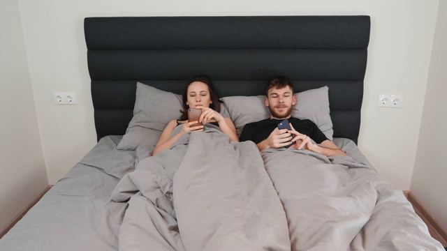 年轻的夫妻正躺在床上，女人正在看丈夫的智能手机。危机的关系。男女躺在床上看智能手机，通过网络交流。视频下载