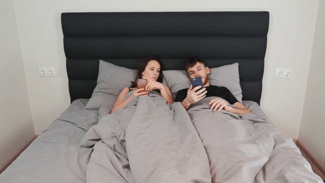 女人和男人躺在床上，手里拿着智能手机。一对年轻的夫妇在灰色的床上看着设备，在网络上发短信。一对男女在卧室里视频下载