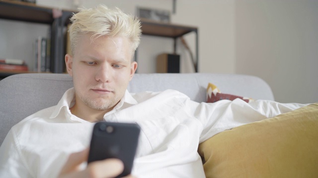 年轻的白人金发男子坐在舒适舒适的沙发上，看着现代的智能手机设备，在里面录音，微笑着和某人聊天。现代通讯方法概念视频素材