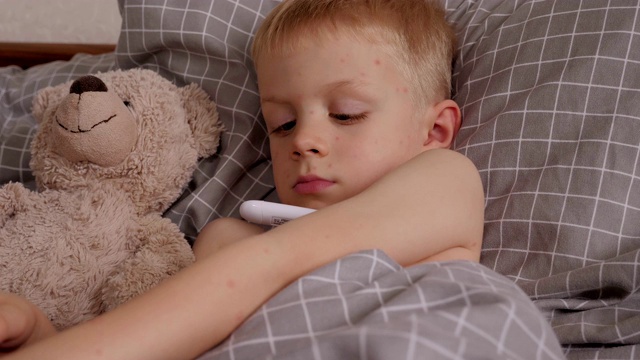 一个生病的小男孩躺在床上，拿着一个温度计和一只泰迪熊。视频下载