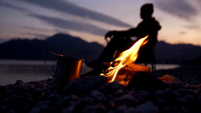 在火旁边放着一个铁杯，一个人躺在海和山的背景下，在地平线上云彩漂浮视频下载