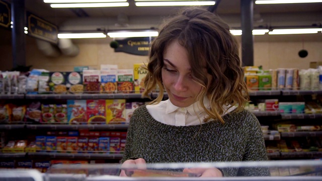 一位年轻的卷发女士在超市里买东西，她正在看产品标签和奶酪营养成分表。架子上的镜头视频素材