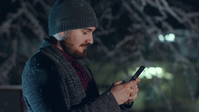 随着雪花从天空飘落，帅气的年轻人正在用手机发短信。股票视频视频素材