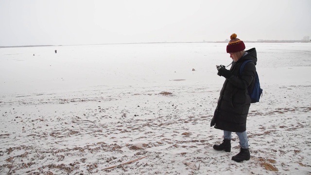 一个女孩在一座桥附近拍摄结冰的河流。女摄影师在冬天拍摄大自然的照片。视频下载