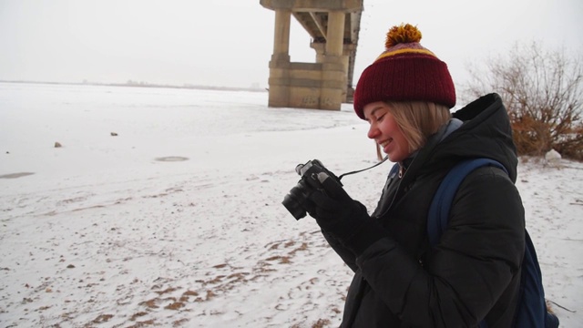 一个女孩在一座桥附近拍摄结冰的河流。女摄影师在冬天拍摄大自然的照片。视频下载