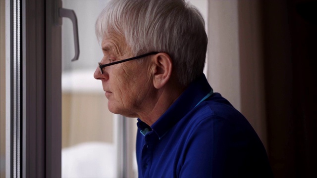 穿着蓝色马球衫，孤独的老人站在窗边。视频素材