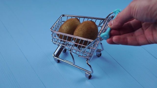 手滚动着一个装饰超市手推车，上面有两个猕猴桃在蓝色的木制背景上视频下载