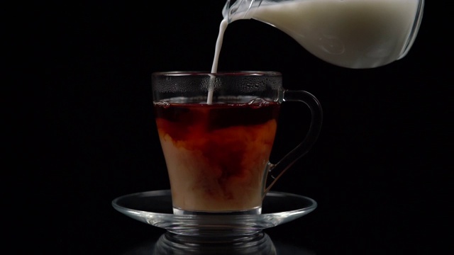 在一个玻璃茶杯和茶托从一个玻璃壶倒牛奶在一个黑色的背景，慢镜头拍摄视频素材