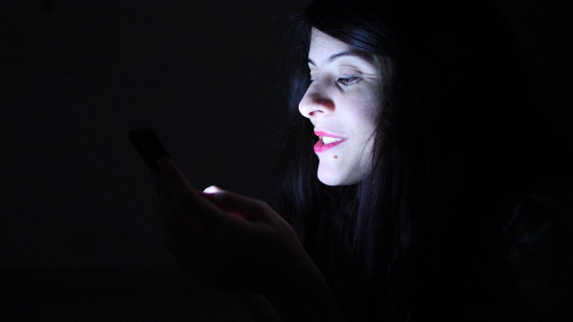 现实生活中的女人笑着看手机设备在晚上在黑暗女孩笑与屏幕设备蓝光辉光视频素材