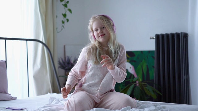 z一代的少女穿着睡衣戴着耳机躺在床上用智能手机听音乐。视频素材