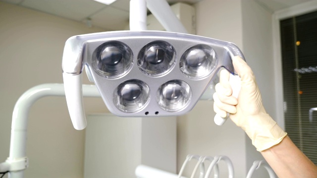 牙医开始治疗，降低牙科灯并打开它。专业的牙医的办公室。特写镜头。充满现代化设备的牙科手术手术室。用手调节头顶的牙科或医疗检查灯。4 k的视频视频素材