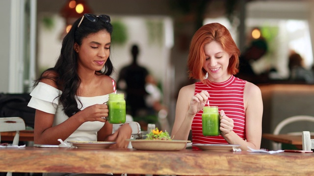 两个不同的漂亮女孩喝着绿色果汁。多文化的年轻女性拿着健康的蔬菜汁视频素材