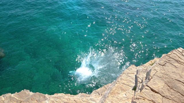 慢镜头:无忧无虑的人从岩石悬崖上跳入深蓝色的大海。视频素材