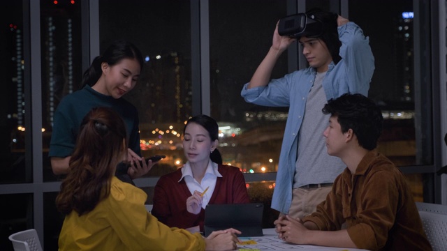 亚洲用户体验开发人员和ui设计师在夜晚的城市灯光下，在现代办公室中使用手机应用原型设计虚拟现实眼镜。创意数字发展机构。移出视频素材