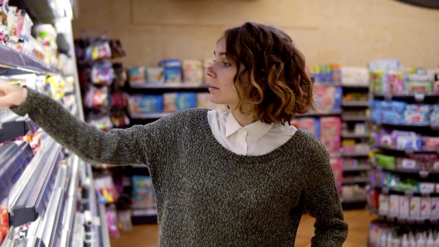 食物，健康理念——一位女士站在超市的冰柜前，选择买一瓶酸奶，拿一瓶放到购物车里。缓慢的运动。近距离视频素材