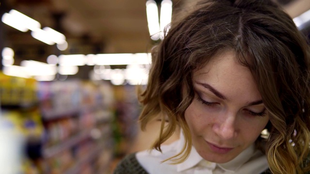 一个年轻的卷发女人在超市购物的特写，她正在阅读一个产品标签和营养事实上的盒子。架子上的镜头视频素材