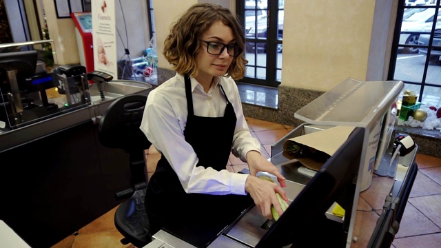 开朗苗条的女售货员穿着白色衬衫和黑色围裙在明亮的超市收银台扫描产品，水果，并把它放入棕色纸袋。高角度镜头视频下载