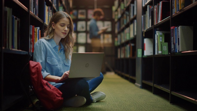 大学图书馆:天才美丽的白人女孩坐在地板上，使用笔记本电脑，为论文写笔记，论文，为课堂作业学习。不同的学生群在学习，为考试而学习。视频下载