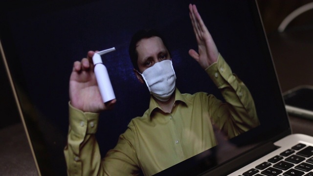 一位戴着医用口罩的视频博主正在用笔记本电脑的网络摄像头录制一段广告视频。他向订户推销喉头喷雾剂。用于预防和治疗疫情期间的病毒感染。视频下载