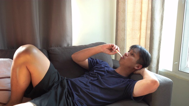 亚洲男人躺在沙发上，用温度计测量家里的温度视频下载
