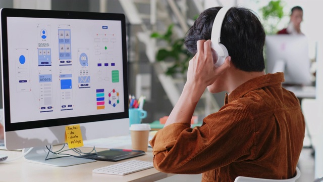 在现代创意办公室，年轻的亚洲程序员男子戴着耳机在电脑桌面上编写移动应用程序，忙碌的亚洲ux, ui设计开发团队，放松的亚洲男性听着音乐工作视频素材