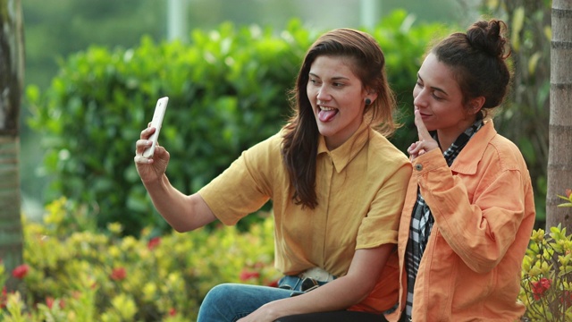 两个朋友在户外用智能手机自拍。千禧一代的年轻女性对着镜头摆姿势视频素材