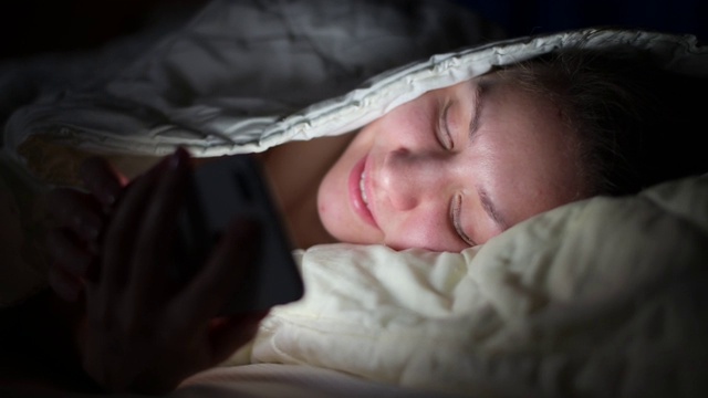 女孩笑着在晚上看手机短信，躺在被窝里视频下载