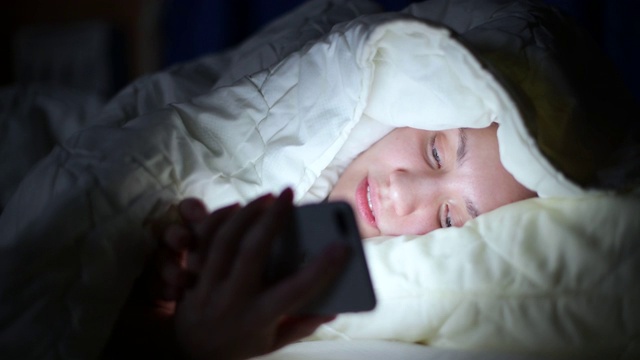 女孩因为失眠正在看社交网络，躺在毯子下面。视频下载