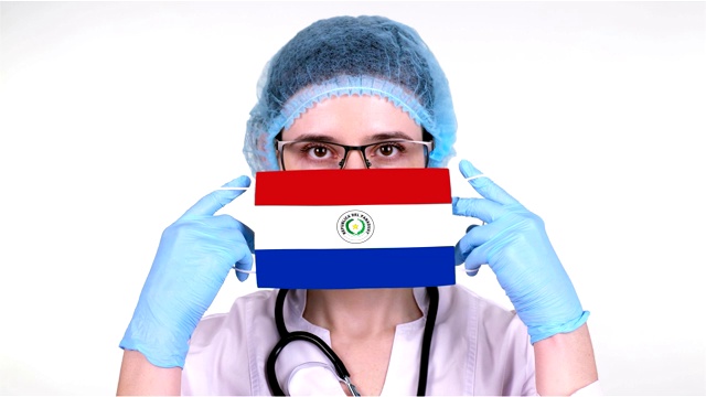 特写镜头。医生戴着眼镜，戴着蓝色医疗帽，双手握着配有巴拉圭国旗的医疗口罩。在冠状病毒全球流行期间，医生护理，国家保护。概念视频下载