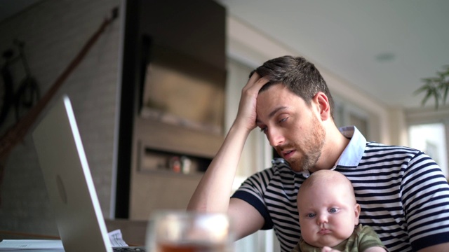 忧心忡忡的男人抱着儿子，在家里用笔记本电脑工作视频下载