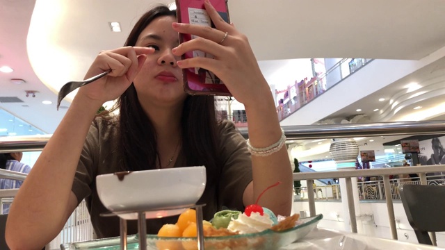 亚洲少女一边在咖啡店吃冰淇淋，一边用智能手机进行网络摄像头，身体状况良好，竖起大拇指视频下载