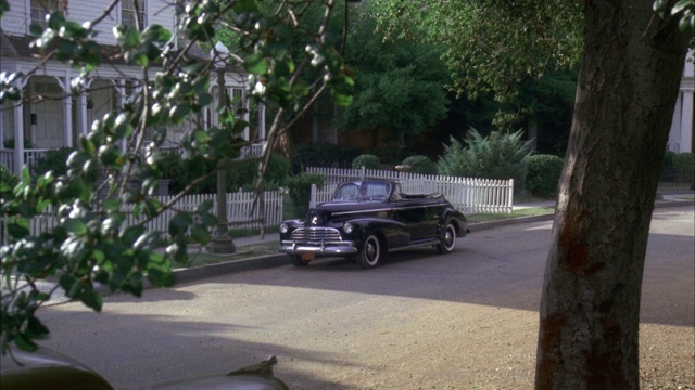 一辆黑色的老爷车从右向右驶去，停在住宅区有白色尖桩栅栏的两层房子前。旧汽车停在街上。街道两旁都是树。视频素材